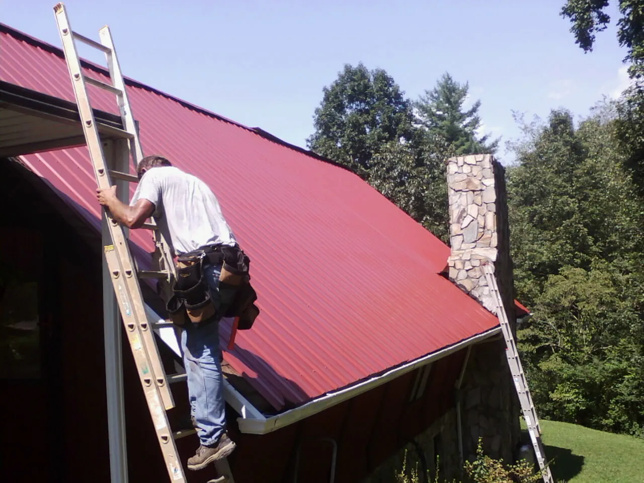 Roofer on a ladder beside a red metal roof and stone chimney — Soffit & Fascia Repair by Roofing Brothers in Le Center